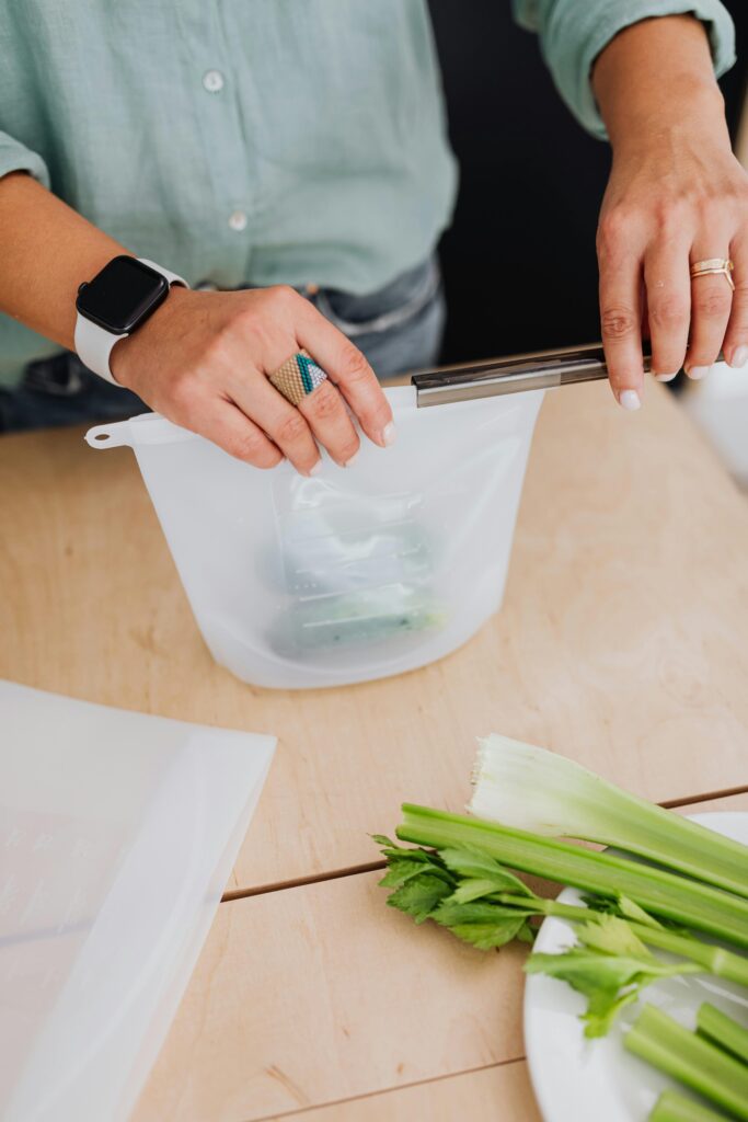 Hands sealing a white silicone reusable food storage bag filled with green vegetables, on a wooden counter with celery, representing leak-proof and sustainable food storage.