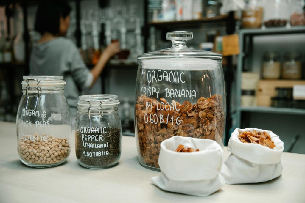 Glass jars and cloth bags filled with organic bulk foods like granola, chickpeas, and spices, showcasing a zero-waste pantry setup.